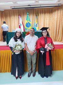 Fotografia de três pessoas em pé, sorrindo. Ao centro, o Diretor-Presidente da FIAM, Otto Narry Tavares da Silva, vestindo camisa social branca. À esquerda, a Reitora da UFOPA, Prof.ª Dra. Aldenize Ruela Xavier, vestindo traje acadêmico com capelo branco e segurando um buquê de flores. À direita, a Vice-Reitora, Prof.ª Dra. Solange Helena Ximenes Rocha, vestindo traje acadêmico com capa vermelha, capelo preto e também segurando flores. Ao fundo, um palco com cortinas douradas e as bandeiras do Pará, do Brasil e do município de Santarém.